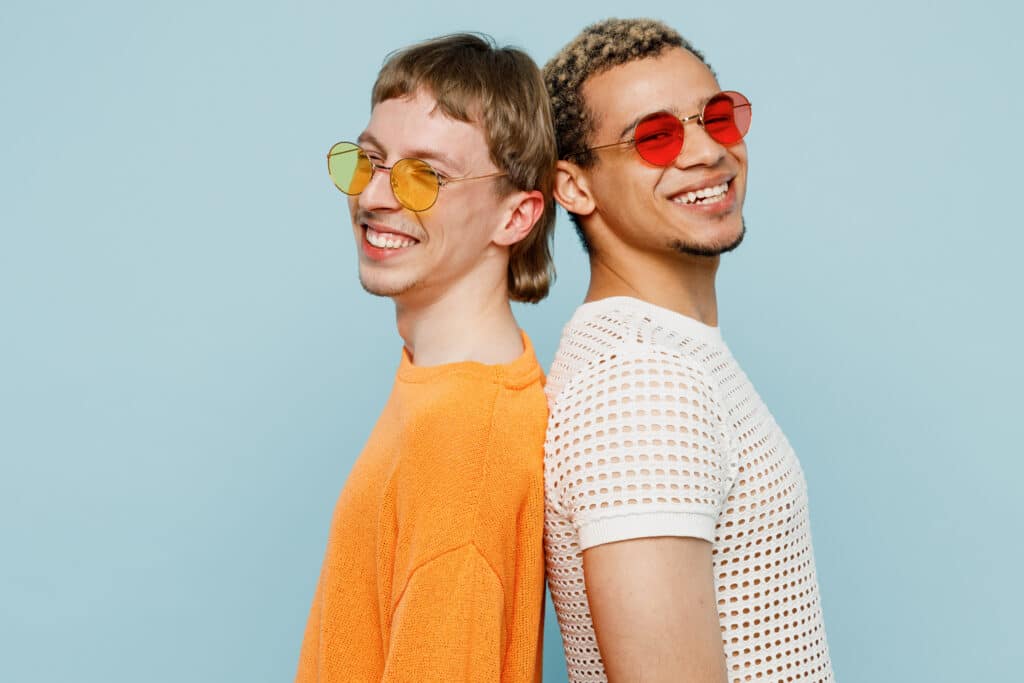 Side view young happy couple two gay men wearing casual clothes sunglasses together stand back to back isolated on plain blue color background studio portrait. Pride day june month love LGBTQ concept.